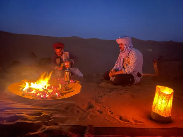 Dos anfitriones del campamento preparan té junto al fuego al anochecer